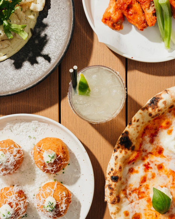 Various plates of food, a pizza and a cocktail in the center